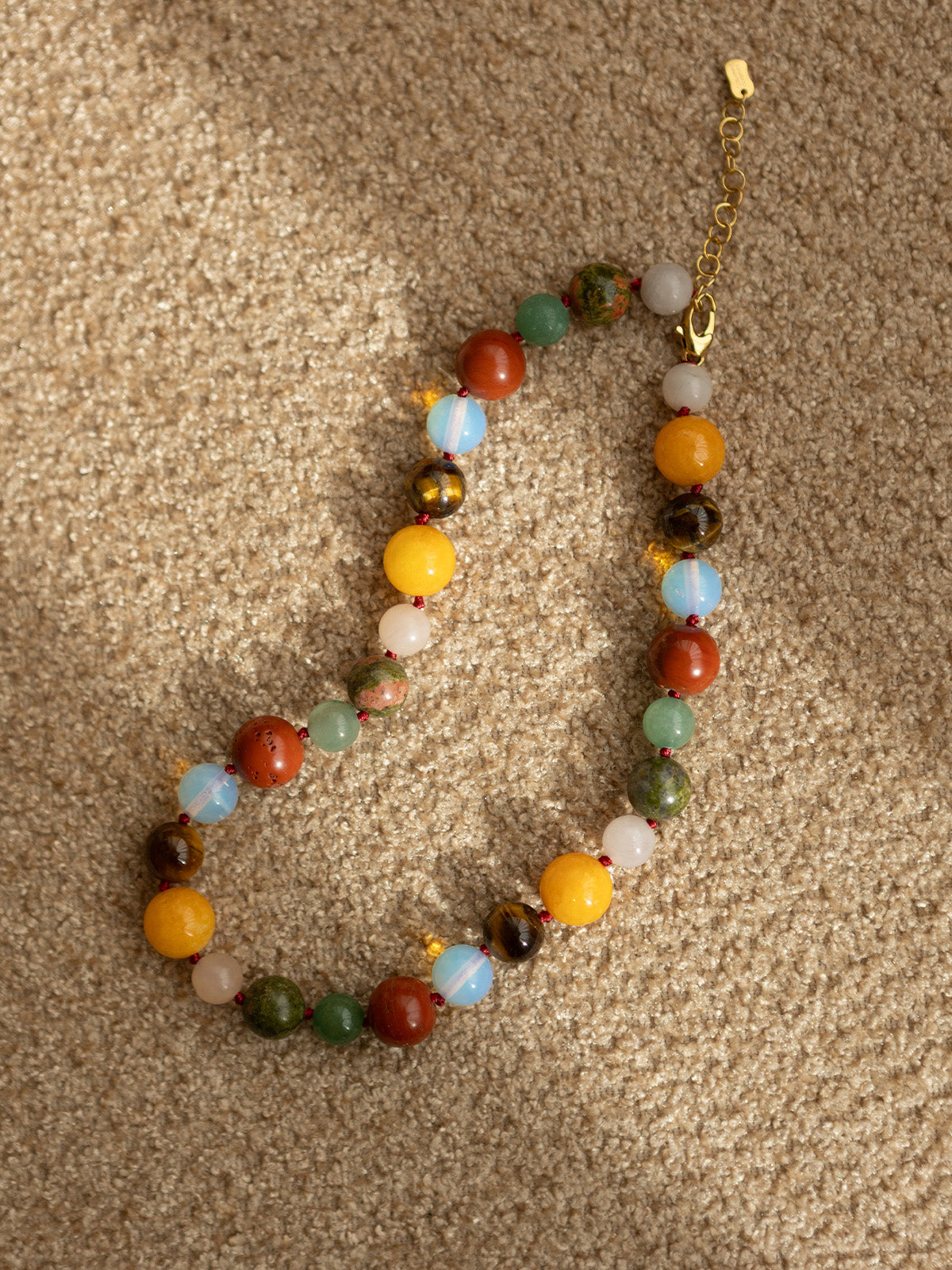 Collana Bold con pietre dure in diaspro rosso, giada gialla, occhio di tigre e opale, fotografia still life ambientata su tessuto beige.
