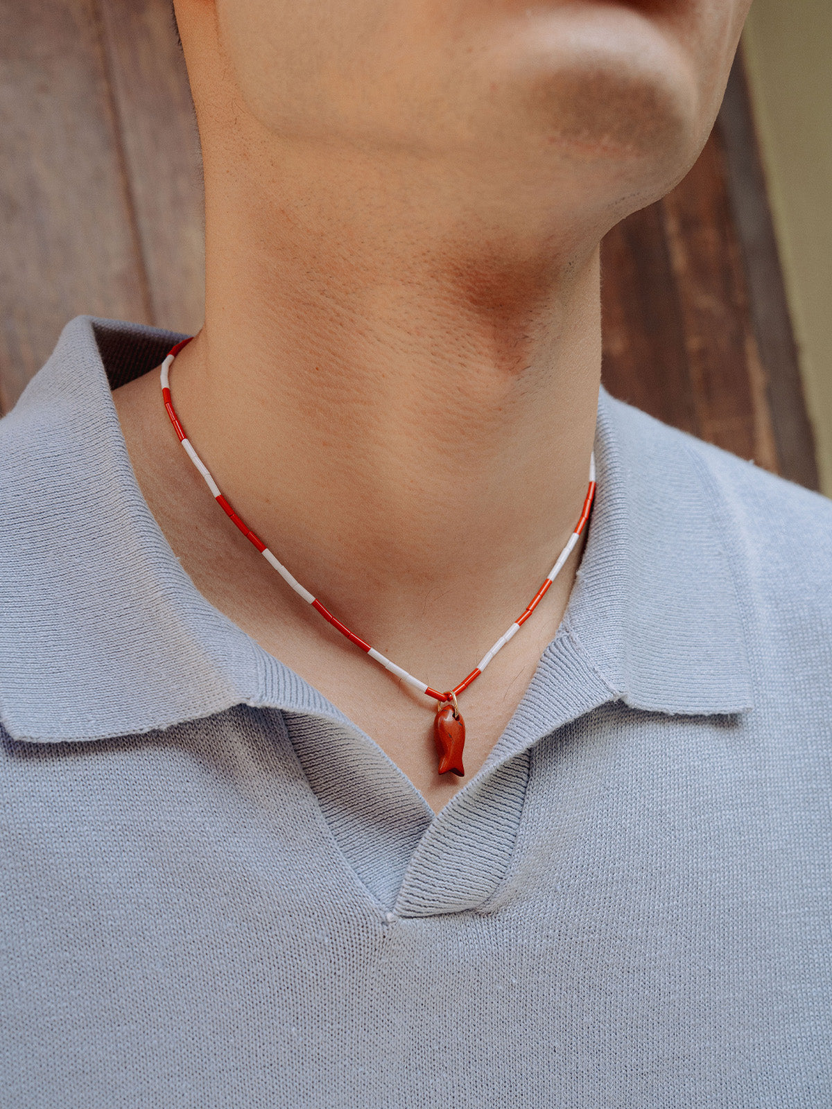 Ragazzo che indossa collanina color block con perline di vetro rosse e bianche e ciondolo pesce in diaspro rosso, fotografato davanti a un muro
