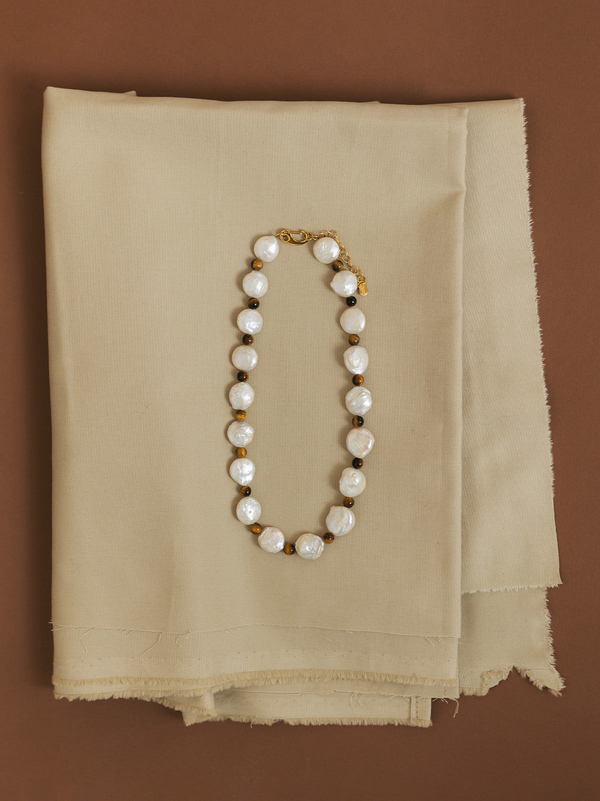 Collanina Bold con occhio di tigre e perle rotonde, fotografia still life creativa su sfondo marrone e tessuto beige.