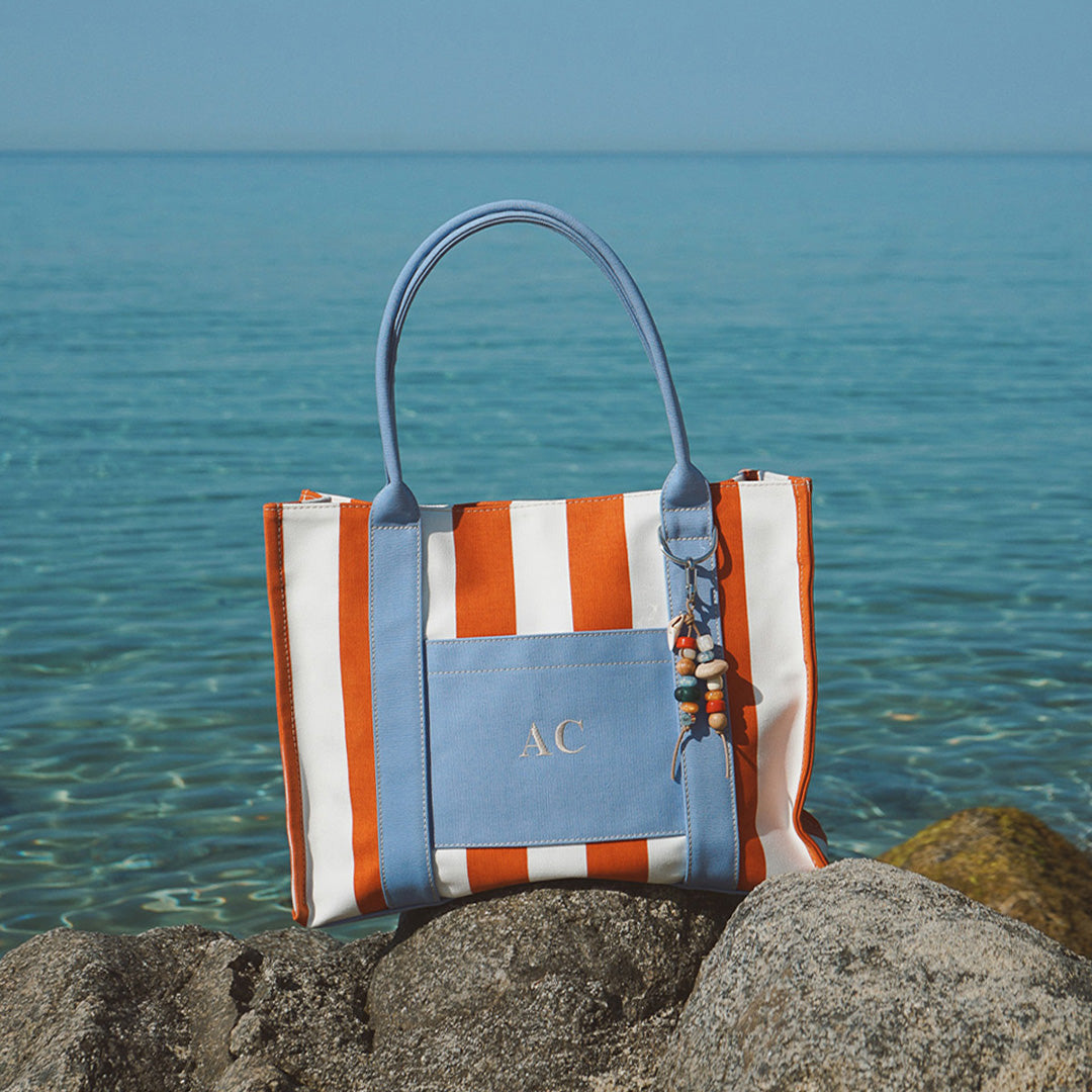 Borsa My Style Bags x Collanine Colorate a righe arancioni e bianche con charm, appoggiata su rocce con vista mare