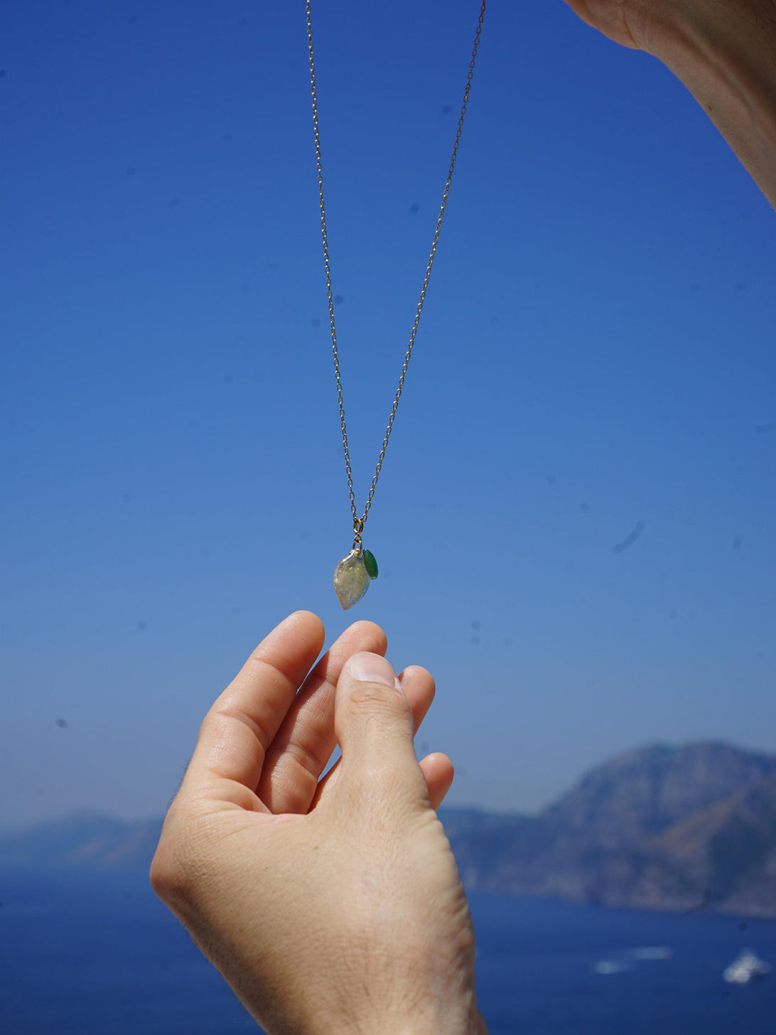 Collana con charm a forma di limone tenuta tra le mani con vista sulla Costiera Amalfitana sullo sfondo
