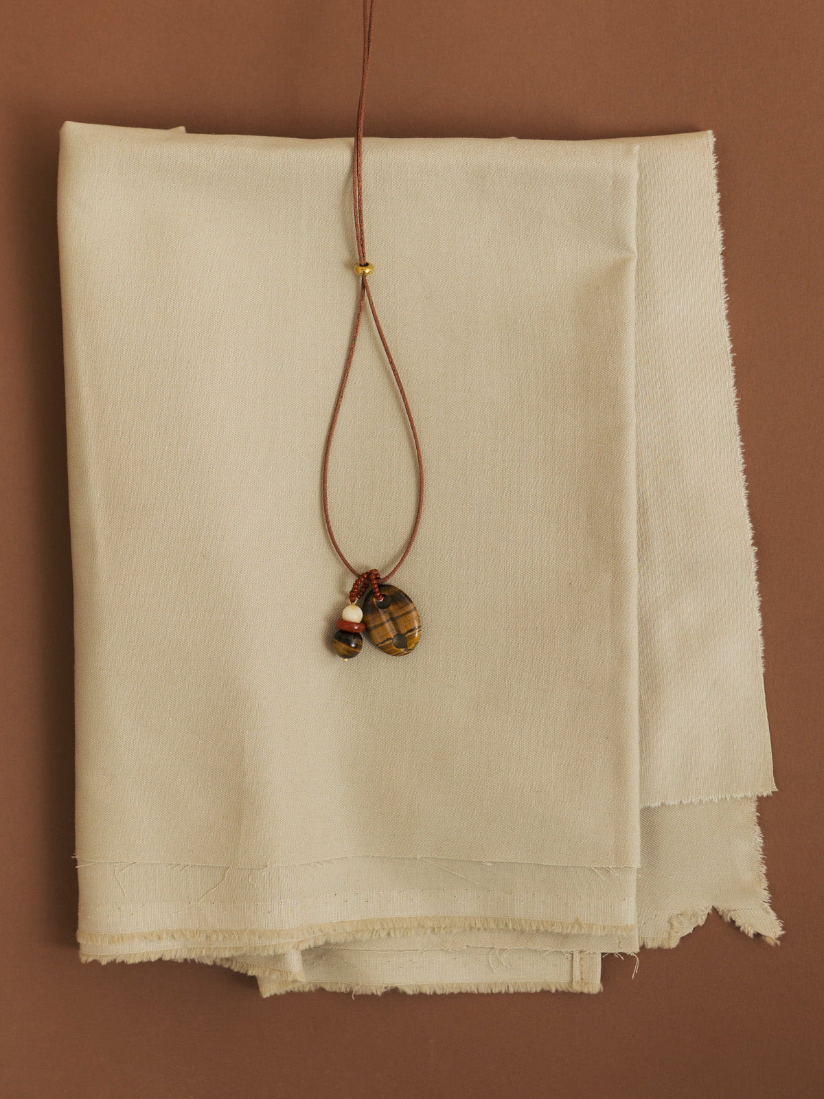 Collana con cordino in raso oro, maglia nautica in occhio di tigre e dettaglio in pietre dure, fotografia still life creativa su sfondo marrone e tessuto beige.