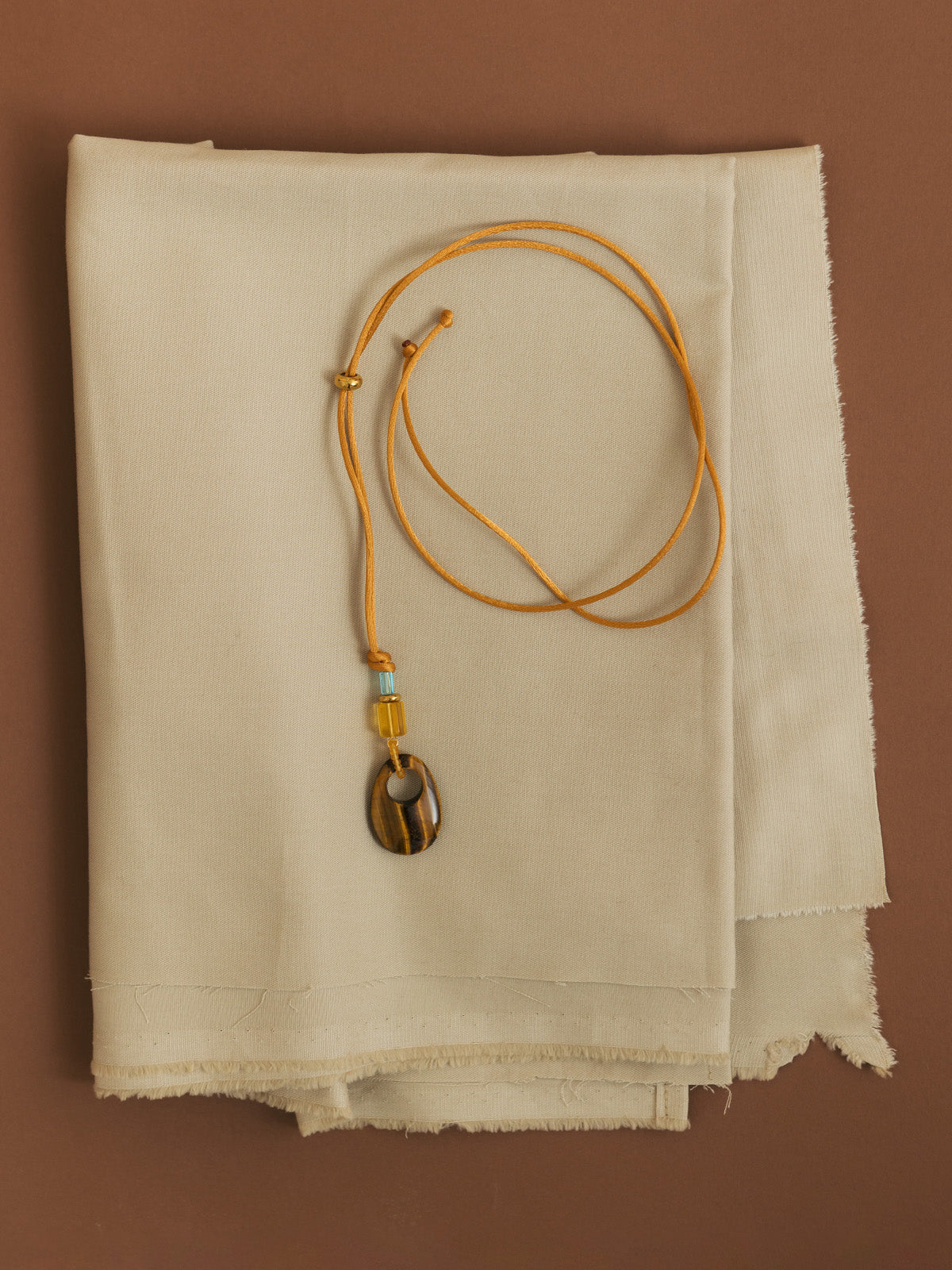 Collana con cordino in raso oro, vetri vintage e occhio di tigre, fotografia still life creativa su sfondo marrone e tessuto beige.